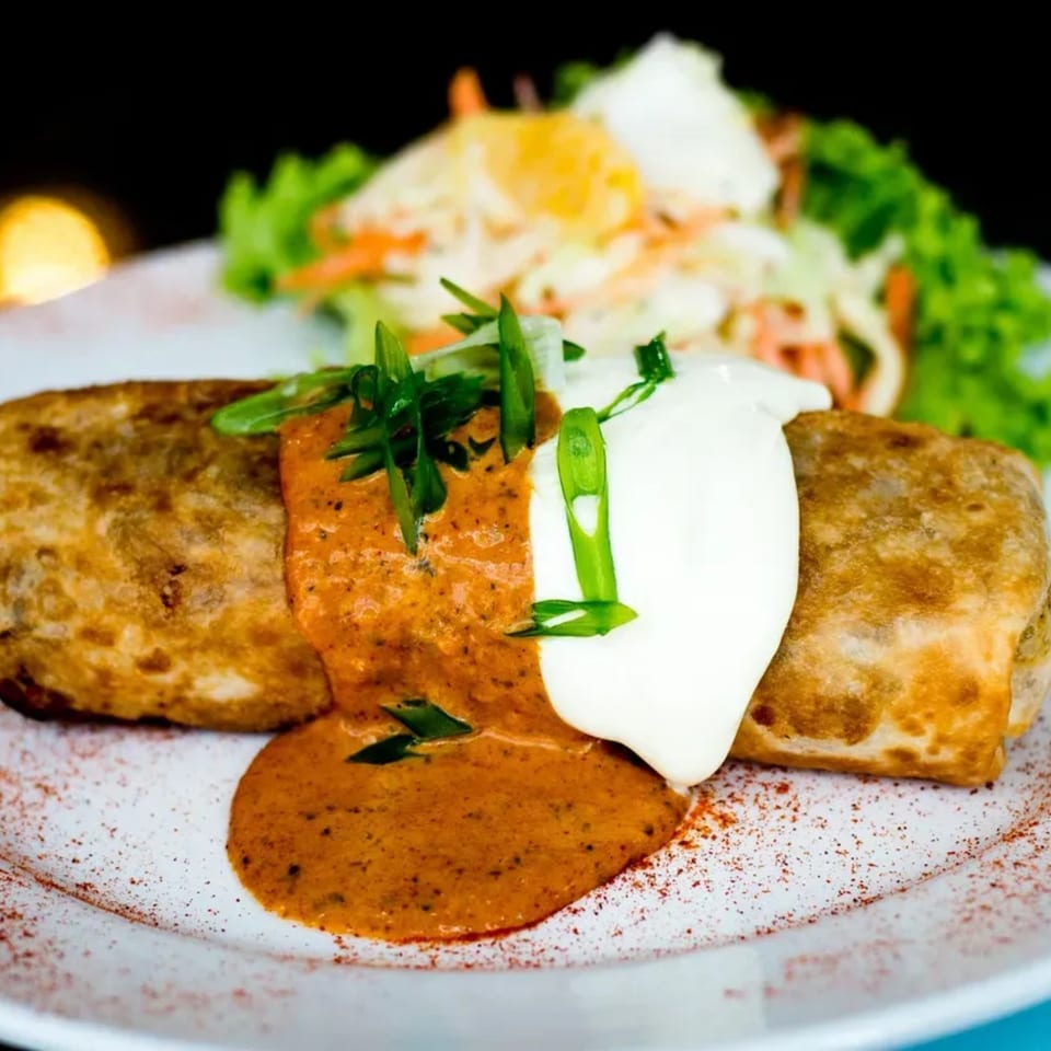 Two golden-brown burritos topped with red sauce, sour cream, and green onions, served with a side salad.