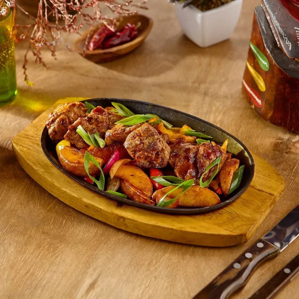 Sizzling meat and potato wedges with green onions and peppers in a black skillet on a wooden board.