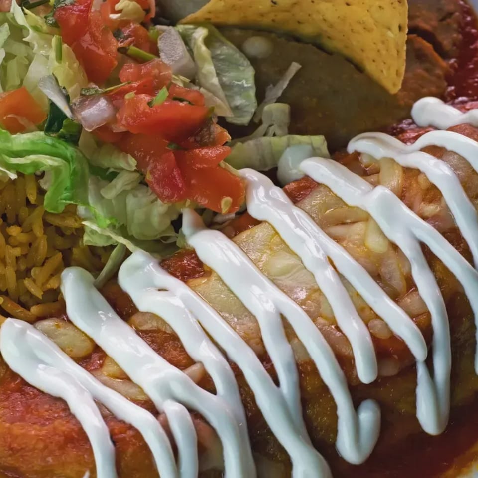 Close-up of enchiladas with sour cream, rice, lettuce, tomatoes, and a tortilla chip.